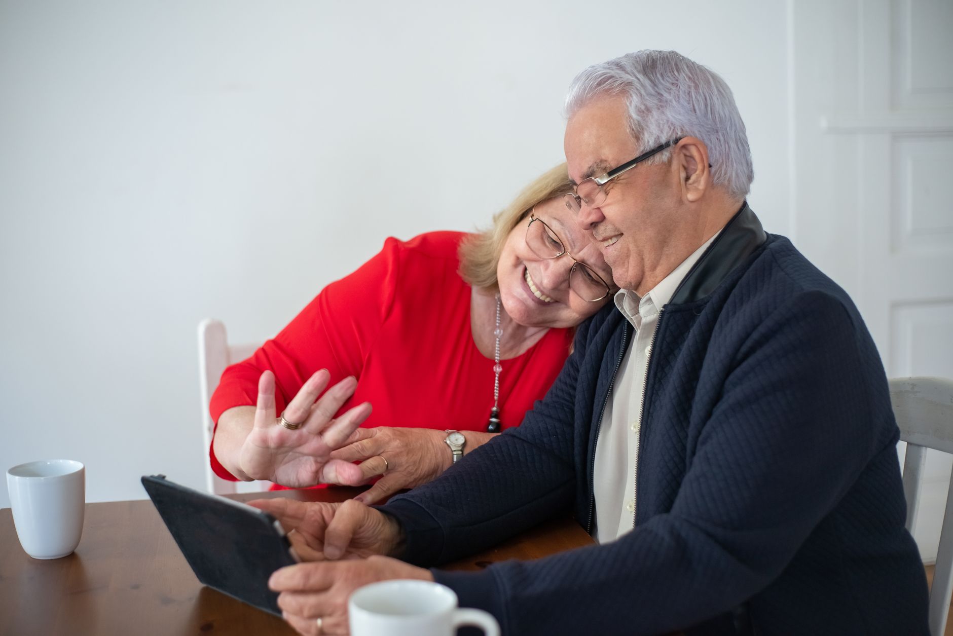 An older adult smiles while using a tablet at a kitchen table with a younger family member sitting nearby.