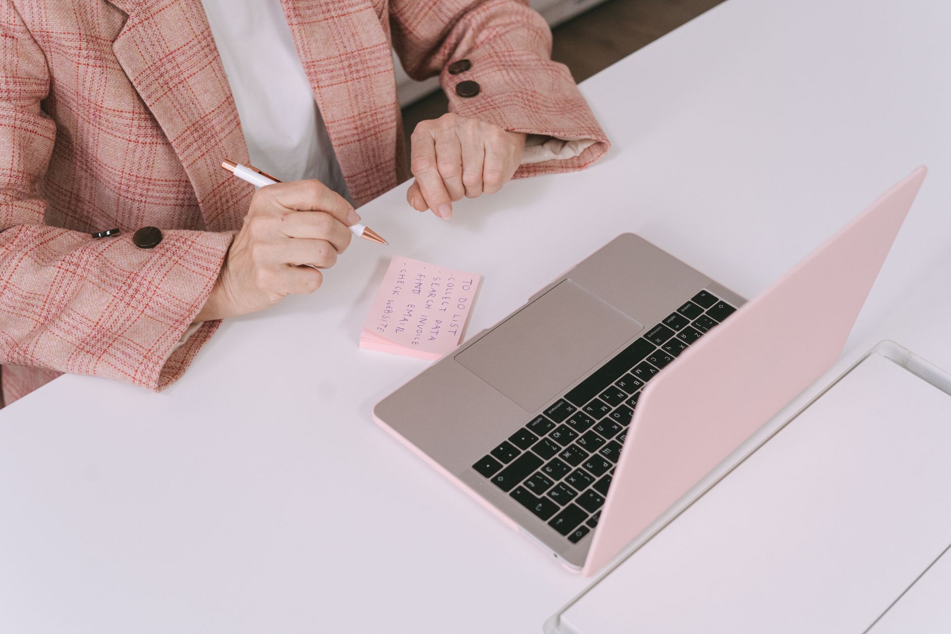 A person writing structured notes beside a laptop at a bright workspace.